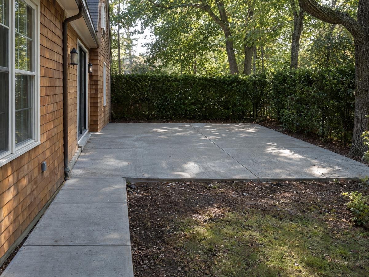 New broom-finished concrete patio and side walkway behind a cedar-shingle house, bordered by a privacy hedge and oaks