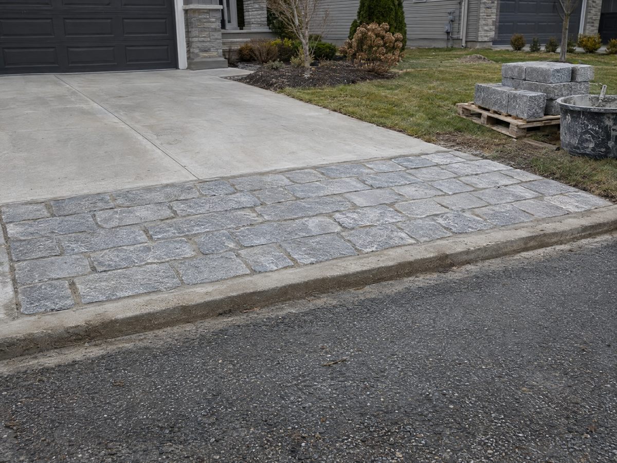 Granite Belgian block driveway apron set in mortar where a concrete driveway meets an asphalt street, spare blocks on a pallet beside a mortar tub