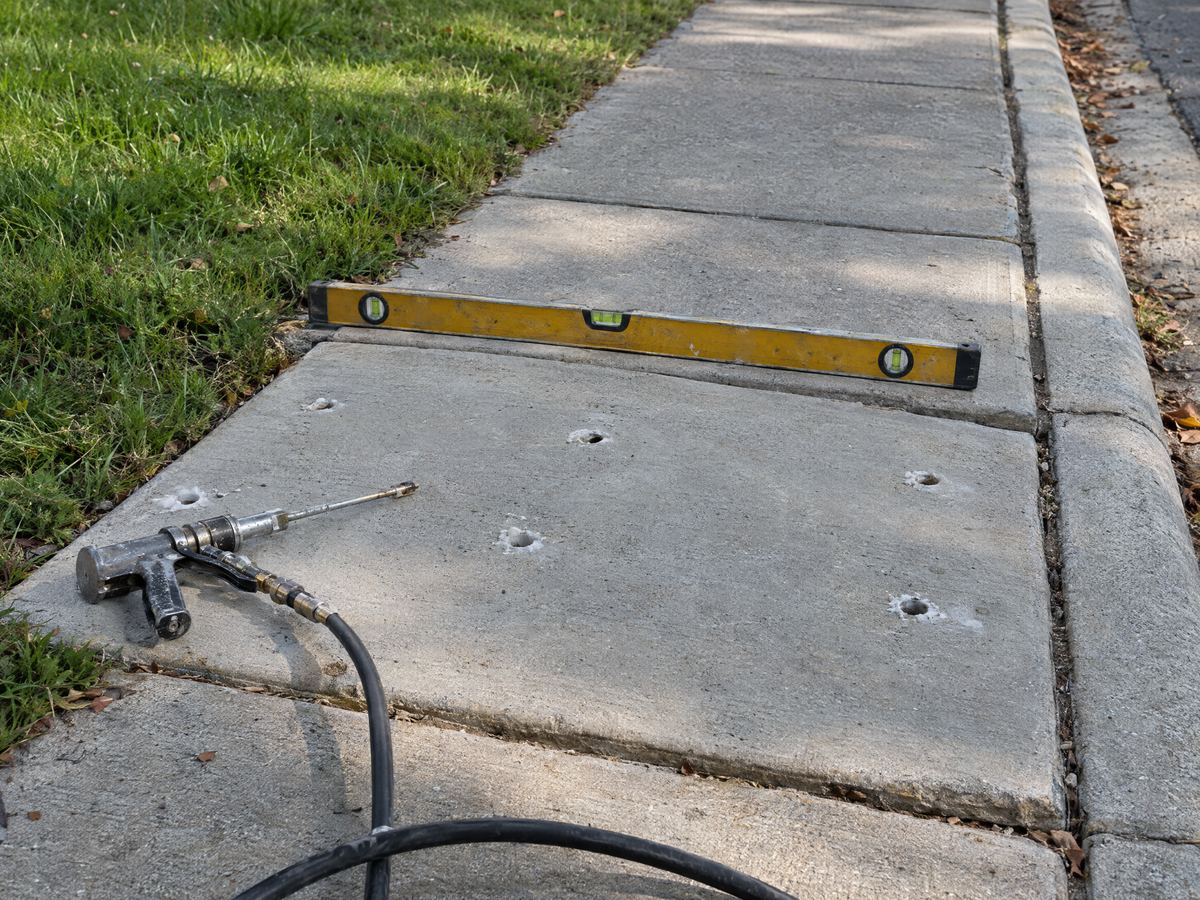Settled sidewalk panel lifted level with its neighbor, patched injection holes, an injection gun and hose and a level across the joint