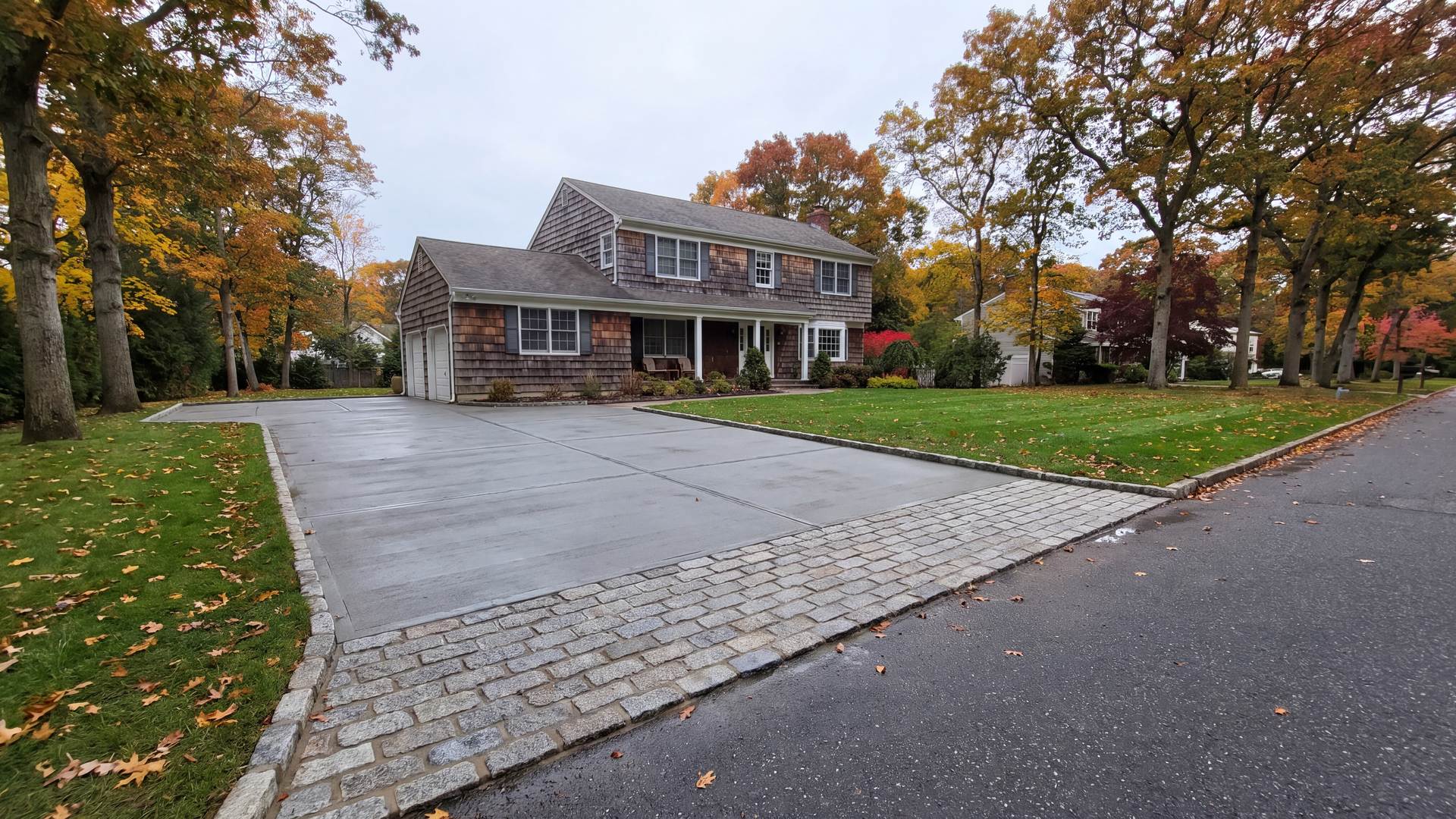 Freshly poured broom-finished concrete driveway with a granite Belgian block apron in front of a cedar-shingled colonial under autumn oaks