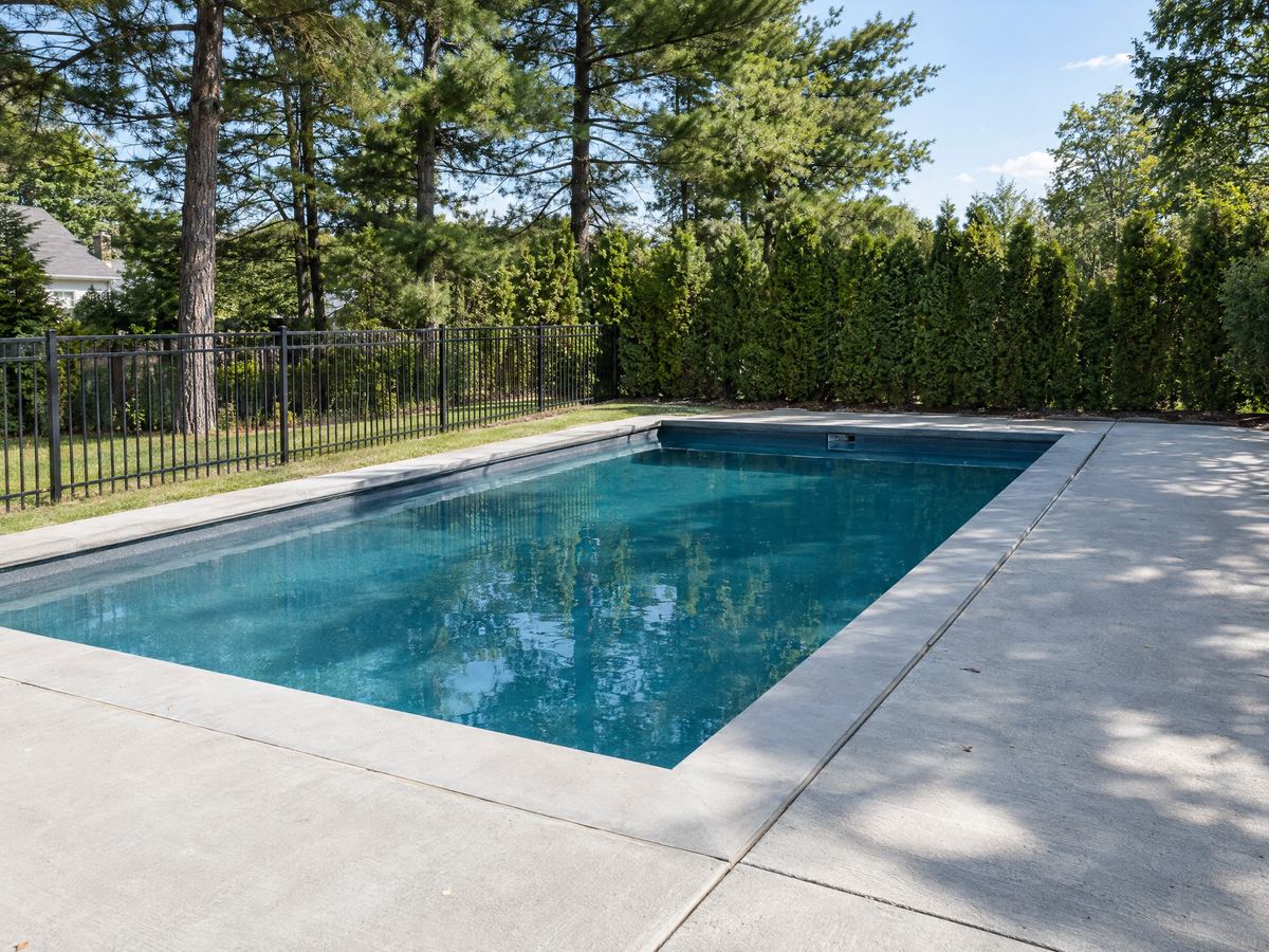Rectangular in-ground pool with a new broom-finished concrete deck, cantilever coping and black aluminum fence among white pines