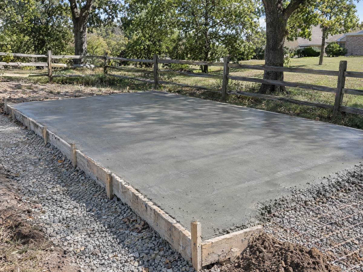 Freshly poured concrete shed pad inside wooden forms with wire mesh and a crushed stone base, split-rail fence and oaks behind